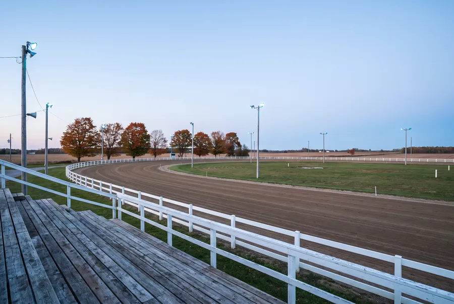 An empty harness racing oval at dusk seen from wooden grandstand benches: a brown dirt track inside a white rail, maples turning orange and farm fields beyond
