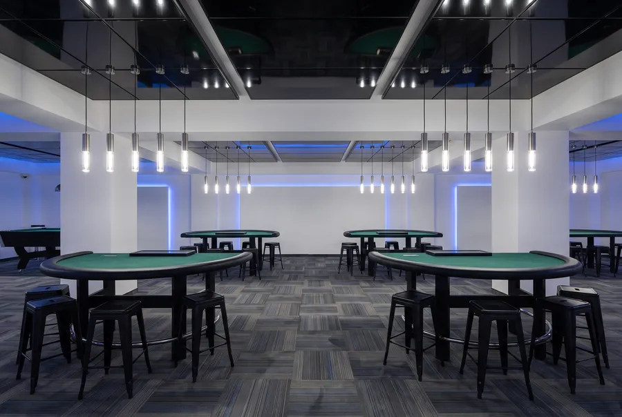 Empty live gaming tables with dark-green felt and black stools under rows of slim pendant lights, before opening